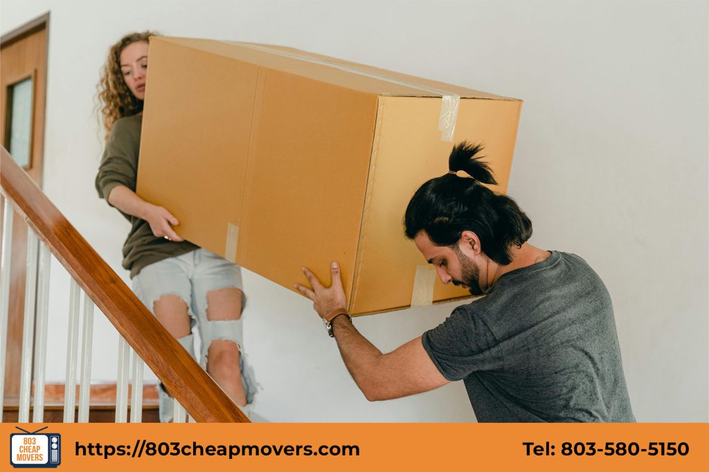 A young couple carries a large cardboard box up a wooden staircase, symbolizing the moving process
